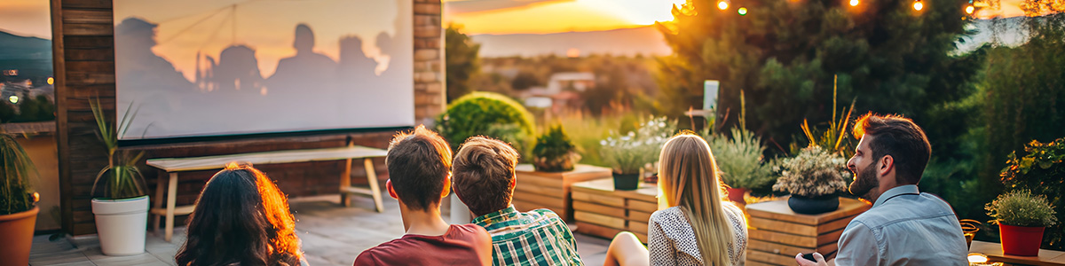 Grupa osób ogląda film na projektorze na tarasie, w tle rośliny, dekoracyjne lampki i zachód słońca.