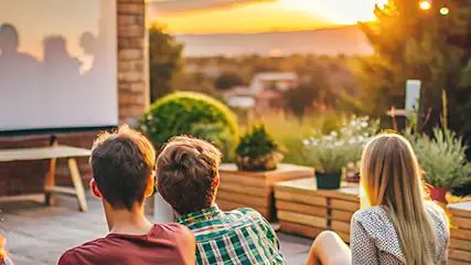 Grupa osób ogląda film na projektorze na tarasie, w tle rośliny, dekoracyjne lampki i zachód słońca.