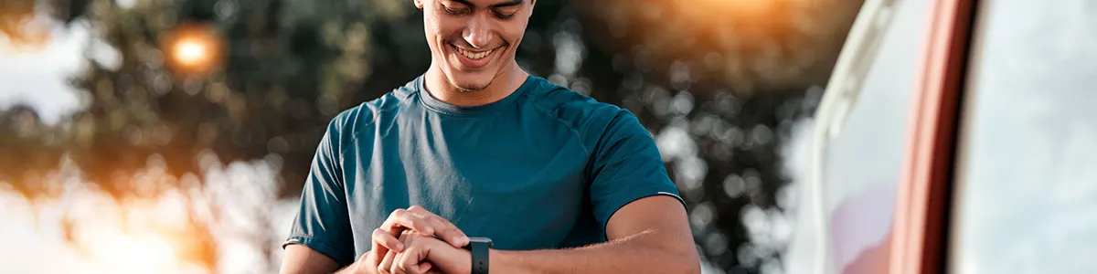 Mężczyzna w niebieskiej koszulce patrzy na czarny smartwatch na swoim nadgarstku. Za nim widoczne są drzewa i jasne światło.