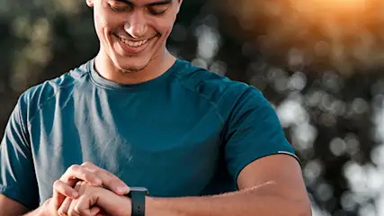 Mężczyzna w niebieskiej koszulce patrzy na czarny smartwatch na swoim nadgarstku. Za nim widoczne są drzewa i jasne światło.