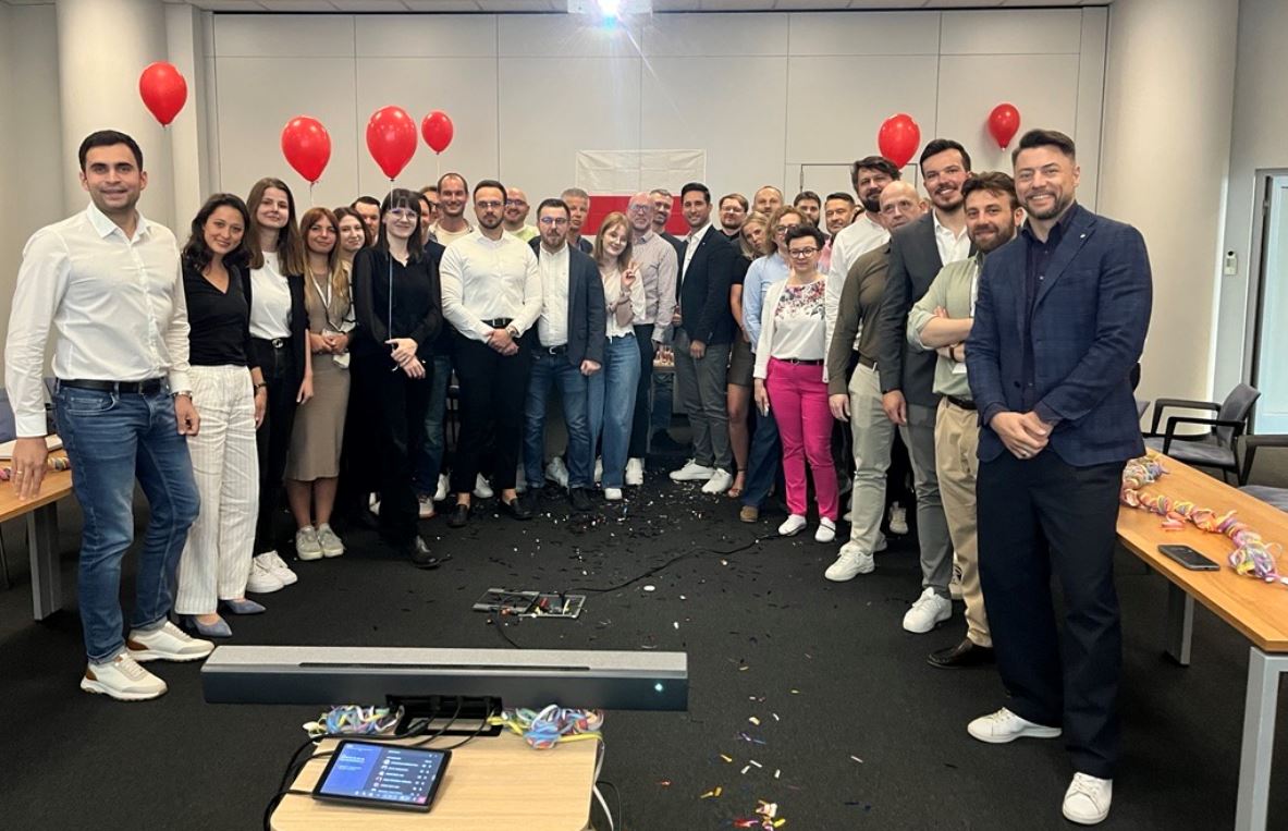 Grupa ludzi stoi w pokoju z czerwonymi balonami i flagą Polski w tle. Na podłodze leżą konfetti.