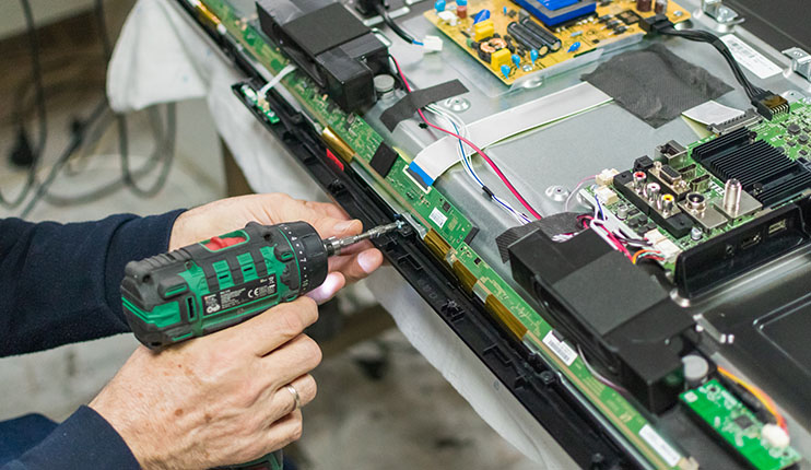 Osoba naprawia telewizor za pomocą wkrętarki. Widoczne są układy elektroniczne i kable.