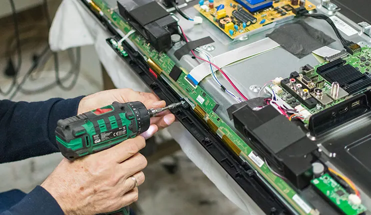 Osoba naprawia telewizor za pomocą wkrętarki. Widoczne są układy elektroniczne i kable.