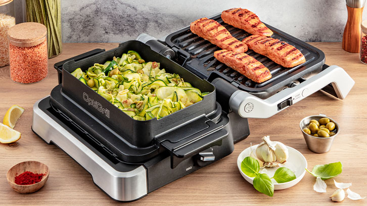 Na stole grill elektryczny z cukinią w pojemniku i czterema kawałkami mięsa. Obok oliwki, czosnek, przyprawy i cytryna.