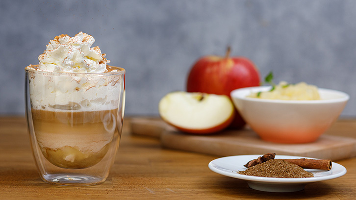 Na stole szklanka z napojem i bitą śmietaną, jabłko, miska z deserem i przyprawy. Tło jest szare, a stół ma drewnianą powierzchnię.