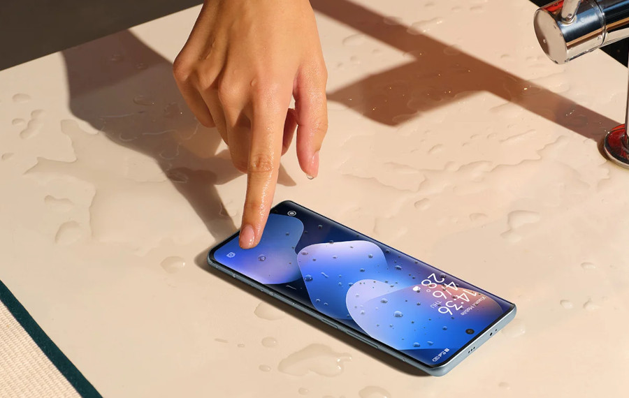 Osoba dotyka smartfona Xiaomi Redmi Note 15 4G, który leży na mokrej, jasnej powierzchni z rozpryskami wody, obok czajnik, w trakcie testowania wodoodporności urządzenia.