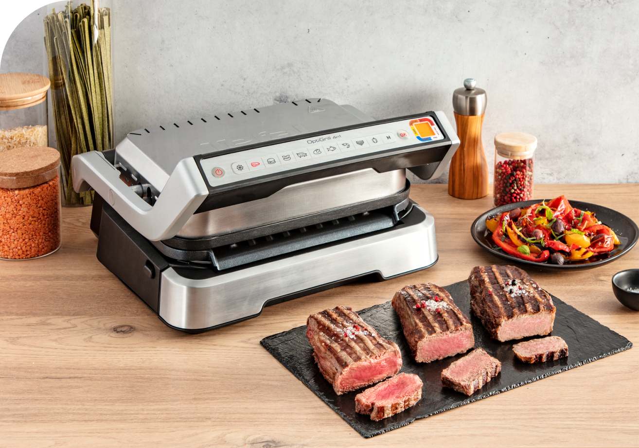 Na drewnianym blacie stoi srebrny grill elektryczny, obok leżą steki na czarnej desce, sałatka i przyprawy.