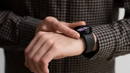 Osoba sprawdza czas na czarnym smartwatchu. Na ekranie widoczna godzina 12:30. Osoba ma na sobie koszulę w brązowo-czarną kratę.