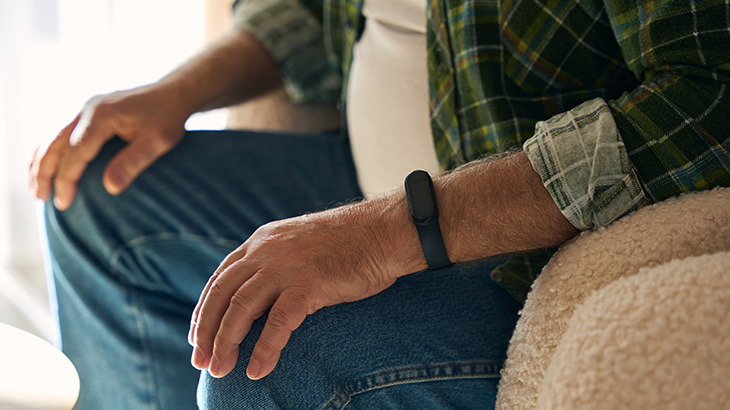 Mężczyzna w dżinsach i koszuli w kratę siedzi, ręce ma oparte na kolanach. Na nadgarstku nosi czarną opaskę. Obok beżowa sofa.