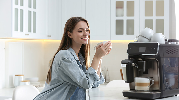 Uśmiechnięta kobieta siedząca obok ekspresu do kawy. W tle białe meble kuchenne.