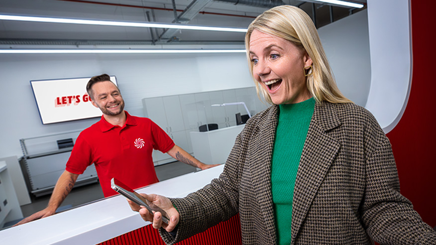 Kobieta trzyma telefon, uśmiecha się. Mężczyzna w czerwonej koszulce za ladą również się uśmiecha. W tle ekran z napisem "LET'S GO".