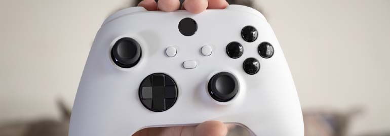 Biały kontroler do gier z czarnymi przyciskami i joystickami trzymany w dłoniach na rozmytym tle.