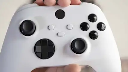 Biały kontroler do gier z czarnymi przyciskami i joystickami trzymany w dłoniach na rozmytym tle.