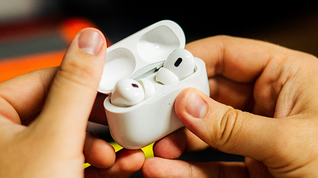 Dłonie trzymają białe etui z otwartym wieczkiem, wewnątrz którego znajdują się białe słuchawki bezprzewodowe.