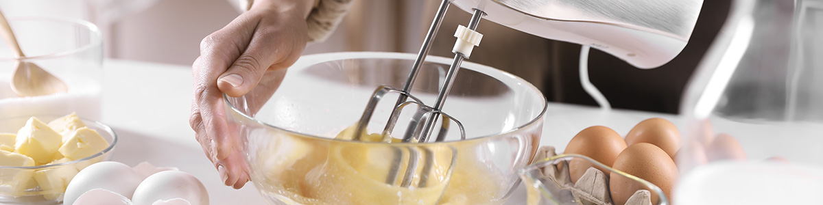 Przezroczysta misa z ciemnożółtym ciastem ubijanym trzepaczkami miksera ręcznego. Obok jajka i masło.