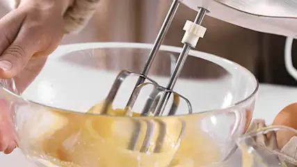 Przezroczysta misa z ciemnożółtym ciastem ubijanym trzepaczkami miksera ręcznego. Obok jajka i masło.