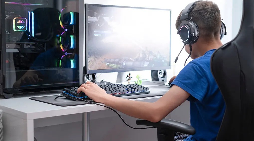 Chłopiec w słuchawkach gra na komputerze; widoczny monitor, klawiatura, mysz, obudowa komputera z podświetleniem i czarne krzesło.