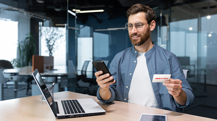 Mężczyzna w okularach trzyma telefon i wizytówkę przy laptopie w biurze.