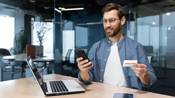 Mężczyzna w okularach trzyma telefon i wizytówkę przy laptopie w biurze.