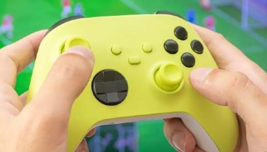 Dłonie trzymają żółty kontroler do gier. W tle widoczny ekran z zielonym boiskiem i postaciami.