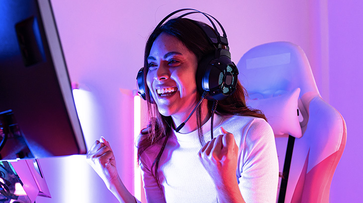 Osoba siedzi w słuchawkach gamingowych przed monitorem. Wykazuje ekspresje zadowolenia, a nawet ekscytacji.