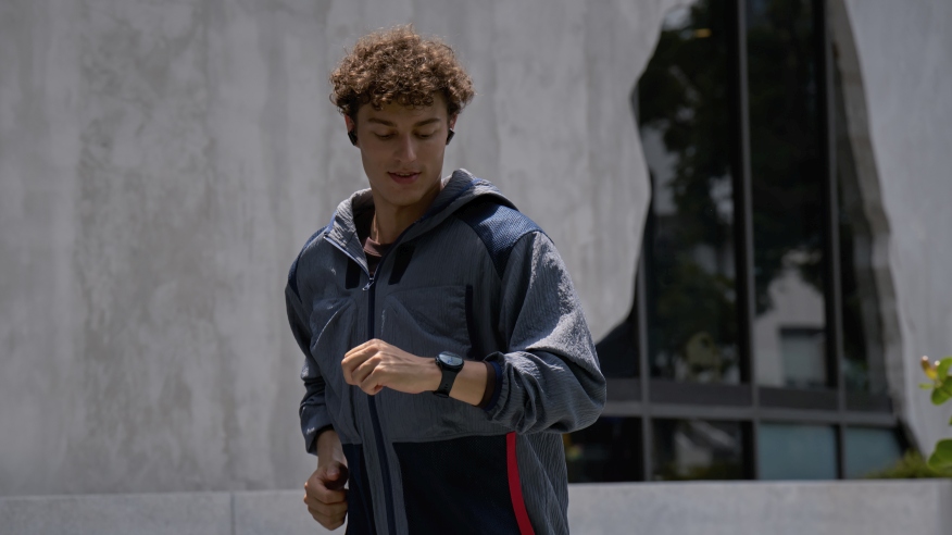 Mężczyzna w szarej kurtce sportowej i czarnym zegarku biegnie na tle budynku z oknami i szarą ścianą.