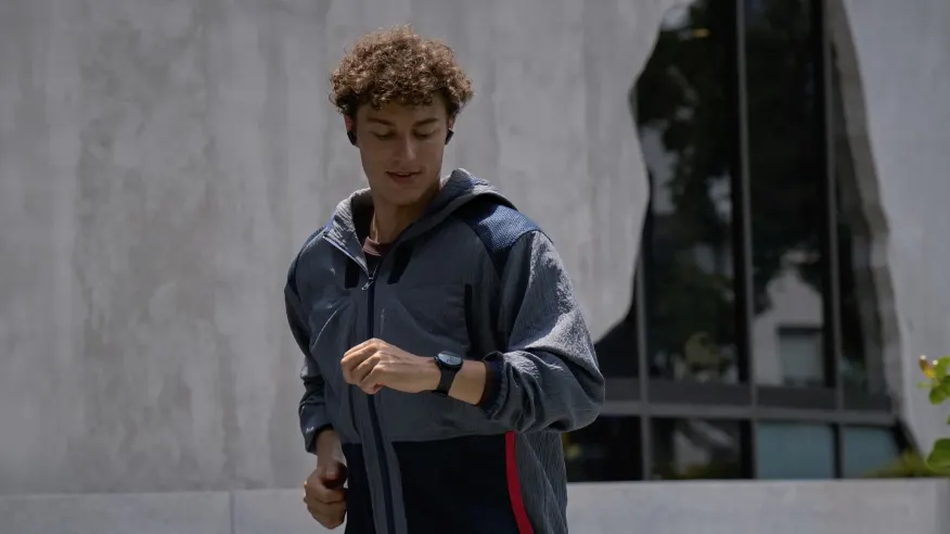 Mężczyzna w szarej kurtce sportowej i czarnym zegarku biegnie na tle budynku z oknami i szarą ścianą.