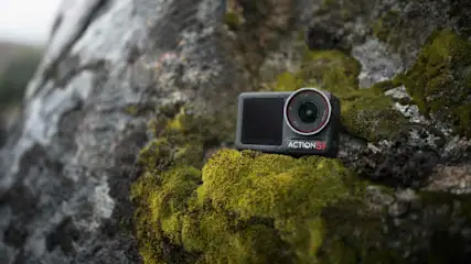 Czarna kamera sportowa leży na mchu porastającym szary kamień. Kamera ma obiektyw z czerwoną obwódką i ekran.