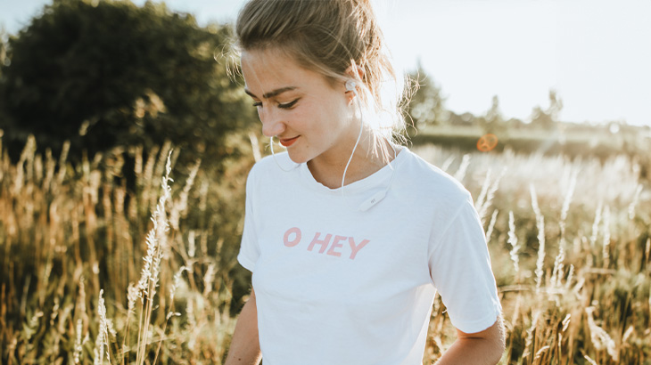 Kobieta w białej koszulce z napisem stoi na łące z wysoką trawą, patrząc w dół.