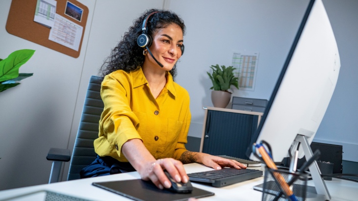 Kobieta w żółtej koszuli i słuchawkach siedzi przy biurku z komputerem i klawiaturą. W tle widać rośliny i tablicę korkową.