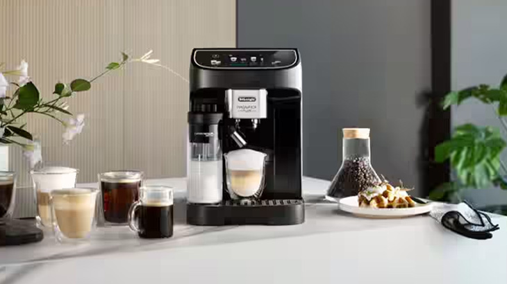 Na stole stoi czarny ekspres do kawy, szklanki z kawą, mlecznik, dzbanek z ziarnami i talerz z ciastkami.