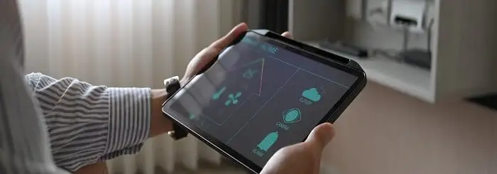 Osoba trzyma tablet z zielonymi ikonami na ekranie. W tle okno i szafka.