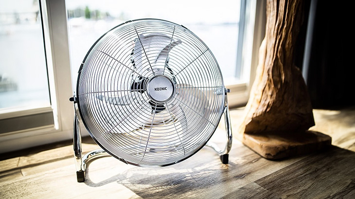 Na podłodze stoi metalowy wentylator z kratką, obok okna i drewniana dekoracja.