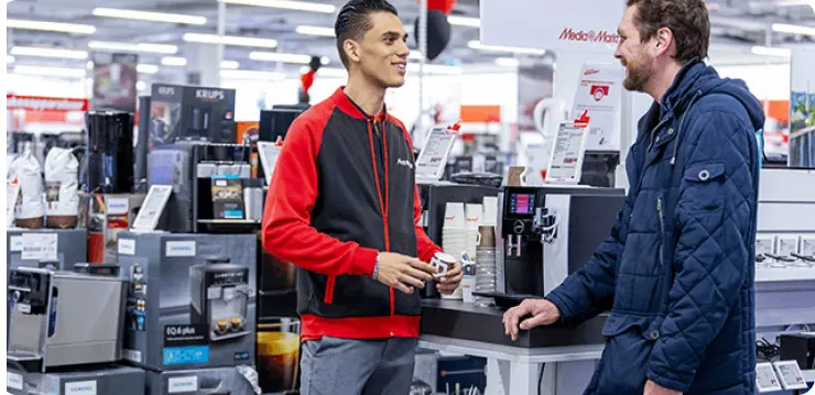 W sklepie, sprzedawca w czerwono-czarnej bluzie rozmawia z klientem w niebieskiej kurtce przy ekspresach do kawy.