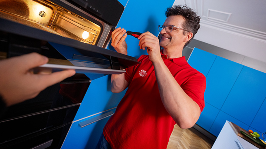 Mężczyzna w czerwonej koszulce naprawia piekarnik w niebieskiej kuchni. Otwarty piekarnik jest oświetlony. Druga osoba podaje tacę.