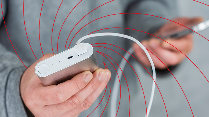 Osoba trzyma srebrny powerbank z podłączonym białym kablem. W tle widać dłoń trzymającą telefon i czerwone linie.