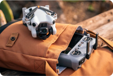 Biały dron leży na brązowym plecaku obok szarego kontrolera, na drewnianym tle.