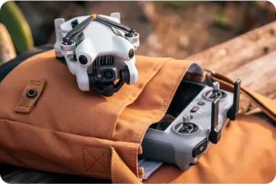 Biały dron leży na brązowym plecaku obok szarego kontrolera, na drewnianym tle.