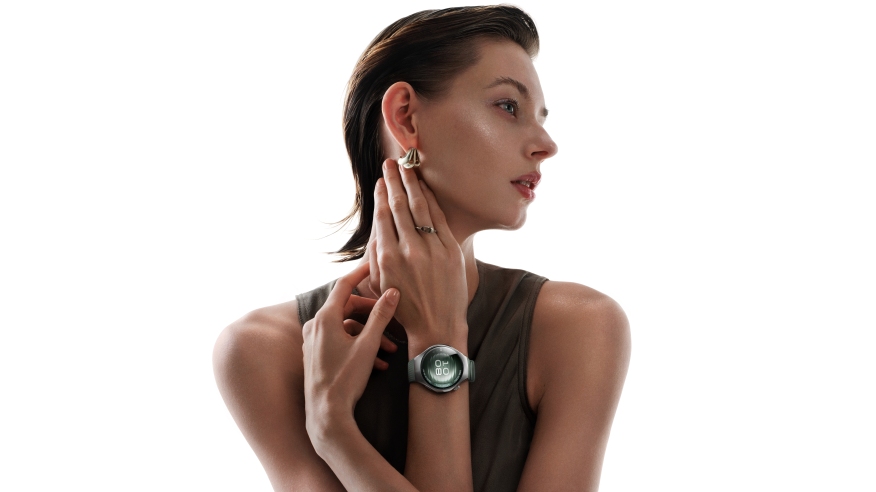 Kobieta z kolczykami i pierścionkiem dotyka szyi. Na nadgarstku ma zegarek. Tło jest białe.