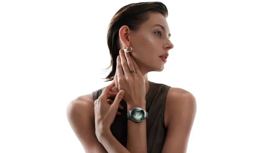 Kobieta z kolczykami i pierścionkiem dotyka szyi. Na nadgarstku ma zegarek. Tło jest białe.