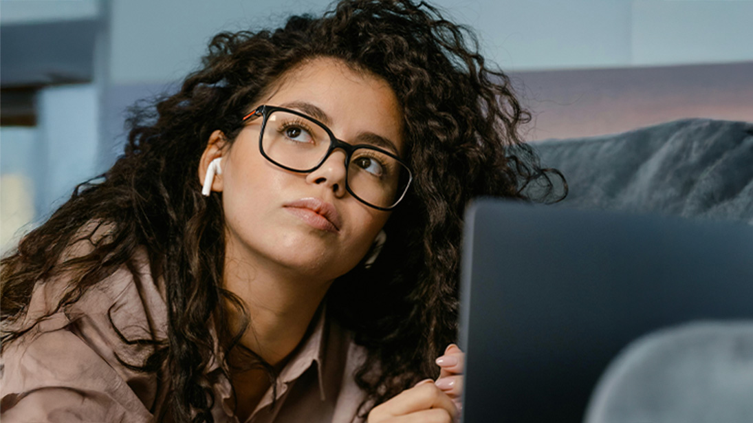 Osoba w okularach i słuchawkach leży na łóżku i patrzy w górę na laptopa. Tło jest częściowo zasłonięte przez szarą pościel.