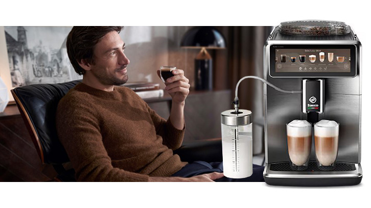 Mężczyzna pije kawę obok ekspresu Saeco, który przygotowuje latte macchiato. Obok widoczny pojemnik na mleko.