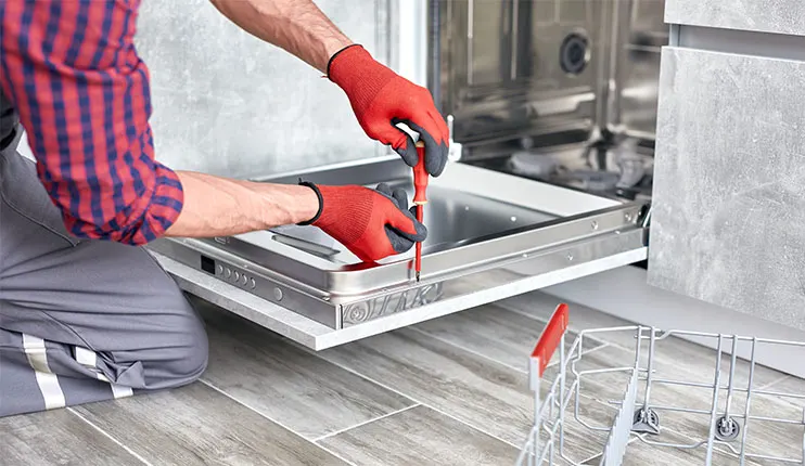 Mężczyzna w czerwonych rękawicach naprawia srebrną zmywarkę na podłodze z paneli. Używa śrubokręta. Obok koszyk na naczynia.