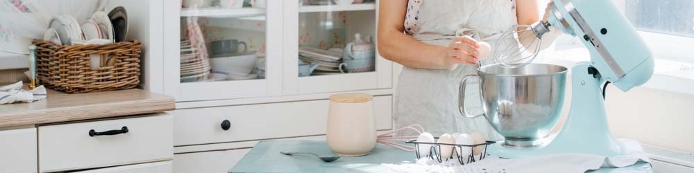 Kobieta w fartuchu używa miksera w kuchni. Na stole misa, jajka, miarka i serwetka. W tle białe szafki z naczyniami.