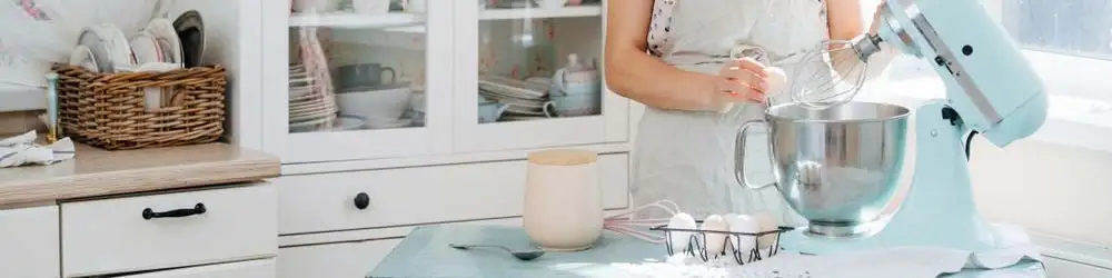Kobieta w fartuchu używa miksera w kuchni. Na stole misa, jajka, miarka i serwetka. W tle białe szafki z naczyniami.