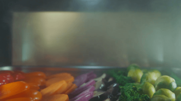 Na pierwszym planie warzywa: marchew, czerwona cebula, brokuły i brukselka. Tło jest rozmyte.