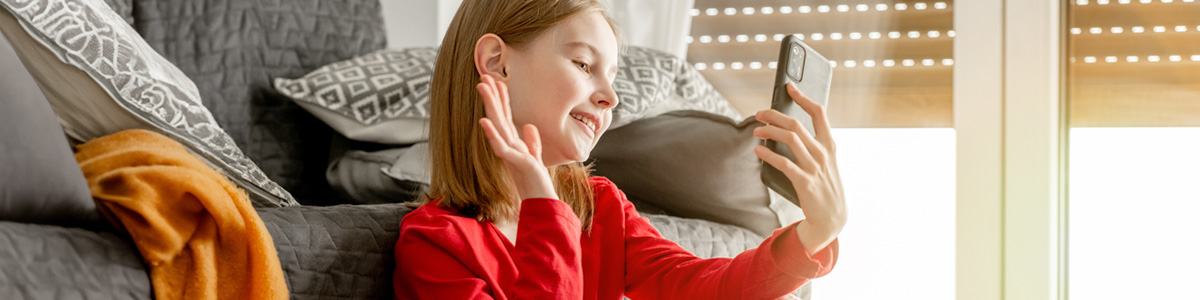 Dziewczyna w czerwonej bluzce macha do telefonu. Za nią szara kanapa z poduszkami i pomarańczowy pled. W tle okno z żaluzjami.