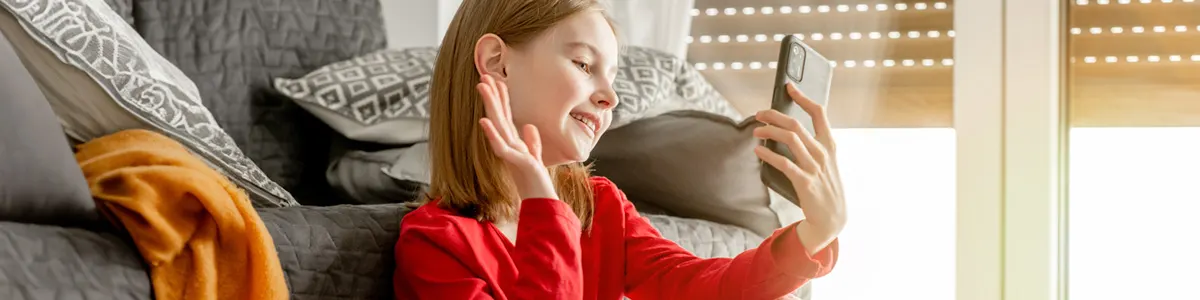 Dziewczyna w czerwonej bluzce macha do telefonu. Za nią szara kanapa z poduszkami i pomarańczowy pled. W tle okno z żaluzjami.