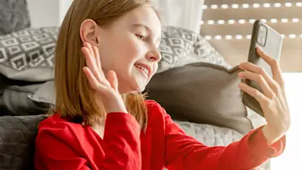 Dziewczyna w czerwonej bluzce macha do telefonu. Za nią szara kanapa z poduszkami i pomarańczowy pled. W tle okno z żaluzjami.