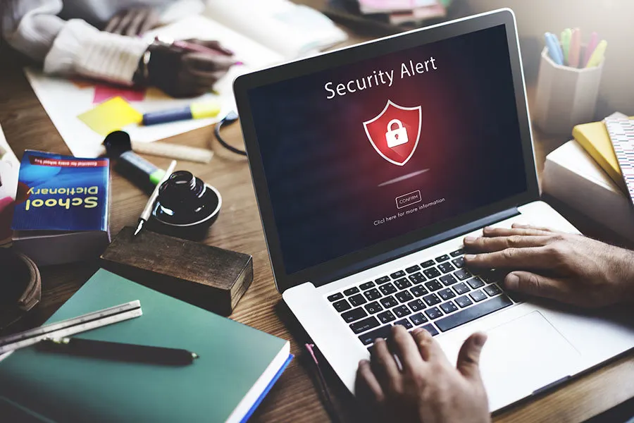 Laptop wyświetla komunikat "Security Alert" z ikoną kłódki. Na biurku leżą książki, notatniki i przybory do pisania. Osoba pisze na klawiaturze.